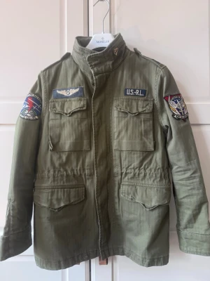 Grön parkajacka från Polo Ralph Lauren - Snygg militärgrön parkajacka från Polo Ralph Lauren med coola patchar på bröstet och ärmarna. Jackan har fyra fickor framtill, hög krage och justerbar midja. Perfekt för dig som gillar streetstyle och utility-vibe.