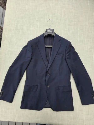 Marinblå modern kostym från Oscar Jacobson - Snygg marinblå kostym från Oscar Jacobson med två knappar framtill, fickor och knappar vid ärmslut. Kavajen har en modern design i slimfit passform och är perfekt för en stilren look. Materialet känns mjukt och följsamt i en blandning av ull, polyester och spandex. Fler bilder med flera mått kan mejlas. Kan skickas eller hämtas. 