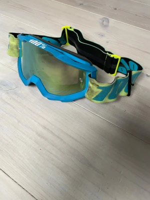 Blåa 100% crossglasögon - Snygga crossglasögon från 100% med blå ram och spegellins. Justerbart band i blått och gult med logga. Perfekta för cykling, motocross eller andra sporter där du vill skydda ögonen mot vind och smuts.