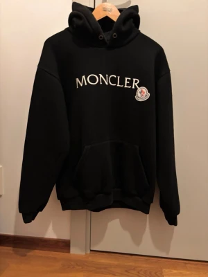 Moncler hoodie - Svart hoodie från Moncler med stor vit logotyp på bröstet och broderad patch på vänster sida. Klassisk känguruficka och huva med snörning. Perfekt för en stilren och trendig look. Strl M. Kom privat vid frågor eller funderingar