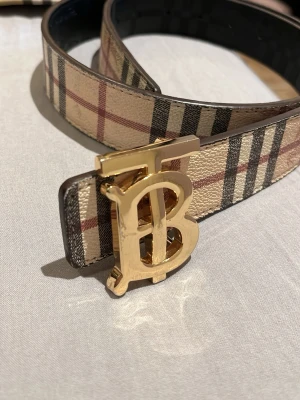 Burberry bälte med guldspänne - Snyggt Burberry bälte med klassiskt rutigt mönster på ena sidan och svart på andra. Bältet har ett stort guldigt TB-spänne och är gjort i skinn. Perfekt accessoar för att lyfta din outfit.