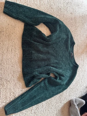 Mörkgrön stickad tröja - Mysig mörkgrön stickad tröja med ribbade muddar och rund hals. Tröjan har en rak passform och är tillverkad i ett mjukt, chenille-liknande material som känns skönt mot huden. Perfekt för lager-på-lager och passar till många olika stilar.