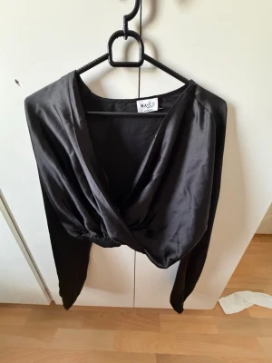 Svart omlottopp i satin från NA-KD - Snygg svart omlottopp från NA-KD i glansigt satinmaterial. Toppen har lång ärm och en croppad passform med djup v-ringning. Perfekt för att lyfta vilken outfit som helst.