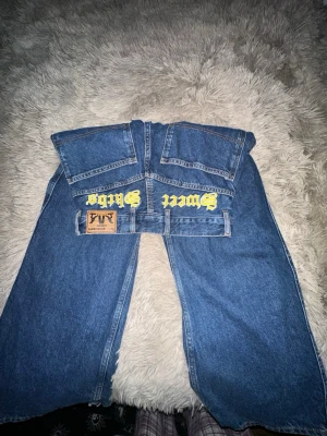 Blå baggy jeans Sweet Sk8rs - Säljer ett par blå baggy jeans från Sweet Sk8rs med stora bakfickor och gul text på baksidan. Jeansen har bred passform och klassisk femficksdesign. Perfekta för dig som gillar streetwear och skön stil.