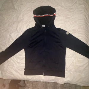Svart Moncler hoodie med randig huva - Svart hoodie från Moncler i storlek M. Tröjan har hel dragkedja, huva med röd och vit randig detalj samt Moncler-logga på ena ärmen. Tillverkad i mjuk bomull, perfekt för chill dagar. Klassisk och stilren design med ribbade muddar.