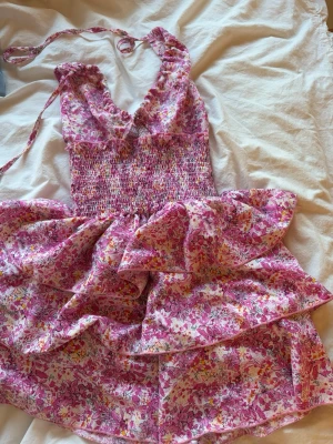 Blommig rosa sommarklänning - Supersöt klänning med rosa och vita blommor, smockad överdel och volangig kjol. Tunna axelband som knyts och djup urringning framtill. Perfekt för varma dagar och ger en romantisk vibe. Tunt, luftigt tyg i bomull.
