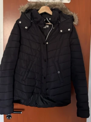Vinterjacka dam - Svart pufferjacka från L.O.G.G. med avtagbar huva och fuskpäls. Jackan har knappar och dragkedja framtill, två snedställda fickor med tryckknappar och quiltad design. Perfekt för kalla vinterdagar och riktigt varm.