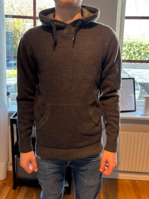 Mörkgrå stickad hoodie - Mysig mörkgrå stickad hoodie med huva och dragsko. Tröjan har en stor magficka och ribbade muddar vid ärmslut och nederkant. Perfekt för en avslappnad stil och passar bra till jeans.