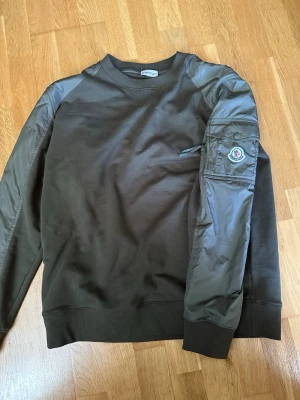 Olivgrön sweatshirt från Moncler - Olivgrön sweatshirt från Moncler med unika nylonpartier på axlar och ärmar. Tröjan har rund halsringning, ribbade muddar och en ficka med Moncler-logga på ena ärmen. Perfekt för en streetwear-look.