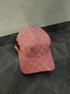 Rosa keps med monogram - Säljer en rosa keps med klassiskt GG-monogram över hela kepsen. Sidorna har gröna och röda ränder som signaturdetalj. Kepsen har böjd skärm och justerbar passform. Perfekt för dig som vill sticka ut med en lyxig accessoar.