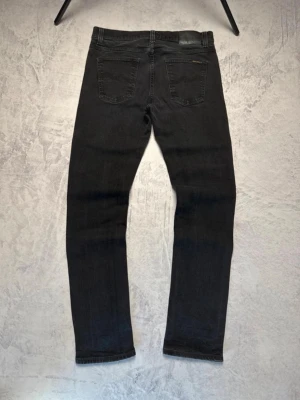 Svarta Nudie Jeans - Säljer ett par svarta jeans från Nudie Jeans med klassisk femficksdesign och raka ben. Jeansen har broderade bakfickor och svart läderpatch i midjan. Perfekta för en stilren och avslappnad look.