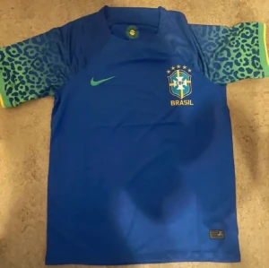 Blå Brasilien Nike matchtröja - Snygg blå Brasilien matchtröja från Nike med leopardmönstrade ärmar i blått och grönt samt gula detaljer. Tröjan har rund hals, korta ärmar och Brasilien-emblem på bröstet. Perfekt för dig som älskar fotboll och vill sticka ut på planen.