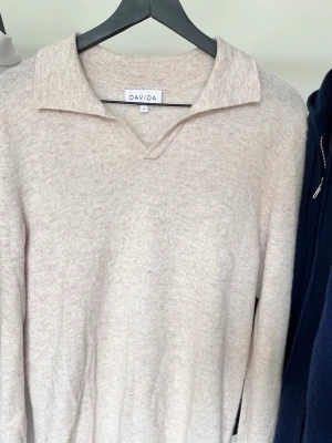 Beige polotröja i kashmir från Davida - Snygg beige polotröja från Davida i mjuk kashmir. Tröjan har klassisk krage med v-ringad öppning, långärmad design. Perfekt för dig som gillar stilrena och bekväma plagg med lyxig känsla. Ny pris är 2399kr