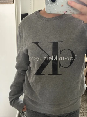 Grå sweatshirt från Calvin Klein - Säljer en grå sweatshirt från Calvin Klein Jeans med stor svart CK-logga framtill. Tröjan har rund halsringning, långa ärmar och ribbade muddar vid ärmslut och nederkant. Perfekt för en avslappnad och stilren look.