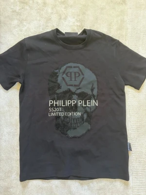 Svart Philipp Plein t-shirt med dödskalle - Cool svart t-shirt från Philipp Plein med en stor dödskalle framtill och texten 'PHILIPP PLEIN SS2011 LIMITED EDITION'. T-shirten har korta ärmar och rund hals. Perfekt för dig som gillar statement-plagg och exklusiv design. Använd 1 gång för att testa bara 