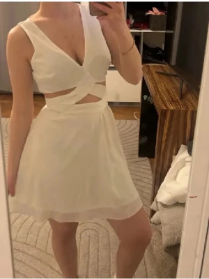 Vit omlottklänning med cut-out - Snygg vit kortklänning med omlott-topp och cut-out-detaljer i midjan. Klänningen är ärmlös och har en v-ringad urringning. Perfekt för sommarens alla tillfällen. Materialet känns lätt och luftigt.