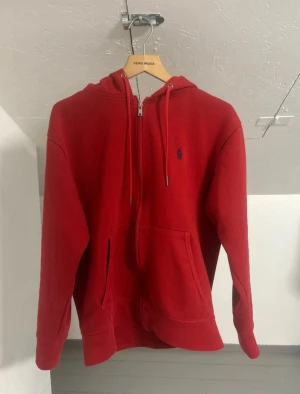 Röd hoodie från Polo Ralph Lauren - Riktigt snygg röd hoodie med dragkedja från Polo Ralph Lauren. Klassisk design med blå broderad logga på bröstet, huva med dragsko och känguruficka. Perfekt för en chill och stilren look.