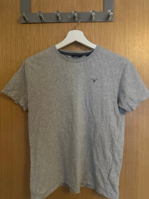 Grå t-shirt från GANT - En stilren grå t-shirt från GANT med liten broderad logga på bröstet. Klassisk rund halsringning och korta ärmar. 158/164 är storleken. Perfekt basplagg i mjuk bomull som passar till det mesta.