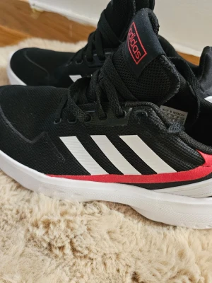 Svarta Adidas sneakers med vita ränder - Svarta sneakers från Adidas med klassiska vita ränder på sidorna och en röd detalj längs sulan. Ovandelen är i meshmaterial för bra andningsförmåga och skon har en vit, chunky sula. Perfekta för en sportig och trendig look.