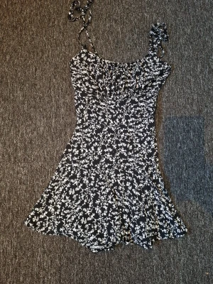 Svart blommig kortklänning - Söt svart kortklänning med vita blommor och smala axelband. Klänningen har en figurnära passform upptill och faller ut i en lätt utställd kjol. Perfekt för varma dagar och enkel att styla med sneakers eller sandaler. Och har inbyggda shorts. 