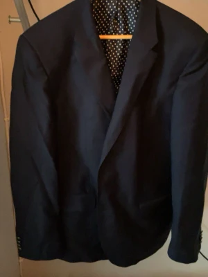 Mörkblå kavaj från Tommy Hilfiger - Snygg mörkblå kavaj från Tommy Hilfiger med klassisk passform. Insidan har ett unikt foder med röda och vita ränder samt prickigt tyg vid ryggen. Perfekt för dig som vill ha en stilren och modern look. Kavajen har knappar vid ärmslut och flera innerfickor.
