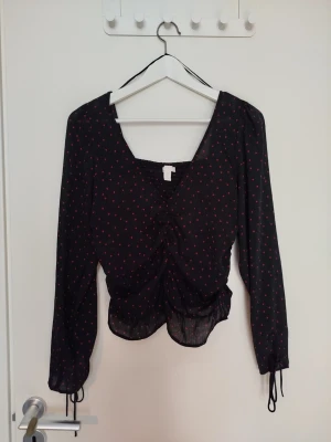 Black blouse with small red hearts - Cute black blouse with small red hearts, a V-neck, and a front tie. Long-sleeved style made of sheer, lightweight fabric with drawstrings at the cuffs. Perfect for those who love romantic details and a trendy look.