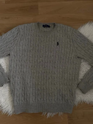 Grå kabelstickad tröja Polo Ralph Lauren - Kabelstickad grå tröja från Polo Ralph Lauren med rund halsringning och klassisk svart broderad logga på bröstet. Tröjan har ribbade muddar vid ärmslut och nederkant och är tillverkad i mjukt material, perfekt för lager på lager.