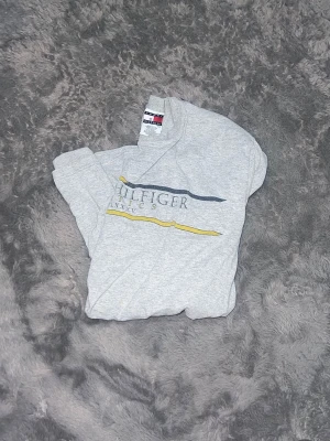 Vintage Tommy Hilfiger Athletics T-shirt -  Snygg vintage t-shirt från Tommy Hilfiger Athletics. Klassisk grå bas med ett schysst retro-tryck i gult och marint på bröstet