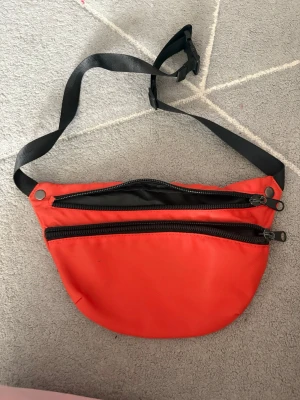 Röd magväska med svarta detaljer - Snygg röd magväska i nylon med två svarta dragkedjor och justerbart svart band. Perfekt för att bära det viktigaste och enkel att ha runt midjan eller över axeln. Väskan har en sportig look och är lätt att matcha till olika outfits.