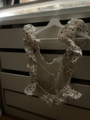 Vit prickig volangtopp med puffärmar - Supersöt vit topp med svarta prickar och stora volangiga puffärmar. Toppen har volangdetaljer både vid axlar och nederkant samt en snygg v-ringning med knyt i ryggen. Perfekt för dig som vill sticka ut lite extra!