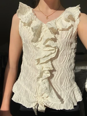 Vit volangtopp med struktur - Supersöt vit topp med volanger längs v-ringningen och framtill. Toppen är ärmlös och har en strukturerad, lite skrynklig yta som ger en cool look. Väldigt elegant och modern.