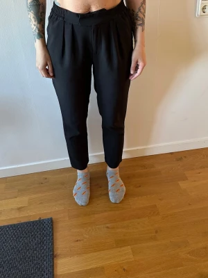 Svarta kostymbyxor med resår - Svarta kostymbyxor med hög midja och resår i sidorna. Byxorna har raka ben, pressveck framtill och diskreta fickor bak. Perfekta för en stilren och clean look. Materialet ser ut att vara en mjuk och följsam textil.