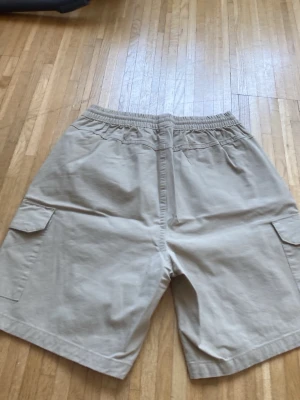 Beige cargoshorts från Adidas - Beiga cargoshorts från Adidas med elastisk midja och två stora fickor på sidorna. Perfekta för en avslappnad stil och har en klassisk Adidas-logga på ena benet. Tillverkade i ett mjukt bomullsmaterial.
