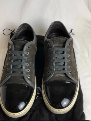 Grå sneakers med glansig tå från Lanvin - Snygga sneakers från Lanvin i grå mocka med grå skosnören och kontrasterande svart lackad tå. Skorna har en vit sula och klassisk låg modell. Perfekta för dig som gillar stilrena detaljer och exklusiv känsla.