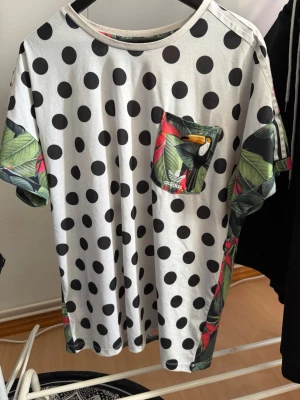 Prickig Adidas t-shirt med bladmönster - Unik t-shirt från Adidas med svartvita prickar och tropiskt bladmönster på ärmar och bröstficka. Fickan har även en liten papegojdetalj och Adidas-logga. Skön passform och tillverkad i mjuk bomull. Perfekt för dig som vill sticka ut lite extra.