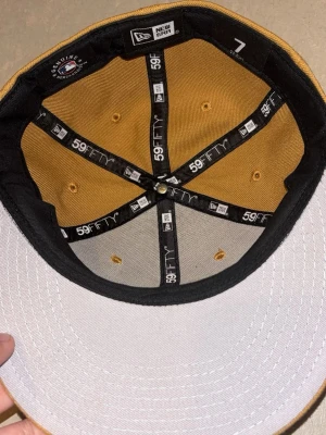 New Era 59FIFTY keps brun/gul - Säljer en New Era 59FIFTY keps i brun/gul färg med svart och vit insida. Klassisk flat brim och broderade ventilationshål. Märkt med 59FIFTY-band på insidan och storlek 7 (55.8 cm). Perfekt för dig som gillar streetwear och snygga accessoarer.