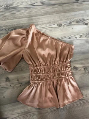 Champagnefärgad oneshoulder-top  - Snygg champagnefärgad oneshoulder-top från Sisters Point i glansigt satinmaterial. Toppen har en puffig ärm, smockad midja och volang nertill. Perfekt för dig som vill sticka ut med en trendig och elegant look.