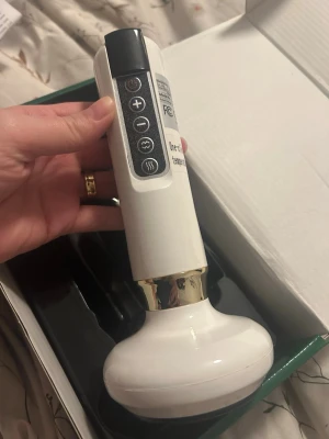 Elektrisk massageapparat vit/guld - Elektrisk massageapparat i vit plast med gulddetaljer. Har flera knappar för olika inställningar och laddas med USB-kabel som medföljer. Ergonomiskt handtag och rundad massageyta, perfekt för avslappning och muskelvård.