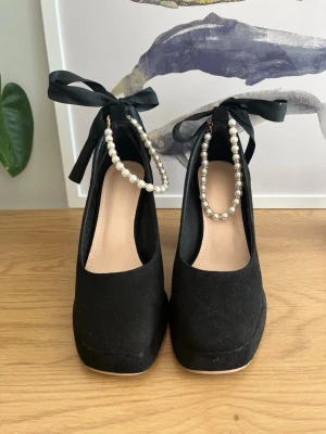 Svarta pumps med pärlband och rosett - Stilrena svarta pumps med chunky klack, dekorerade med pärlband runt vristen och stora svarta rosetter. Skorna har rund tå och är tillverkade i mockaliknande material. Perfekta för dig som vill sticka ut med en trendig och elegant look. I storlek 36. Från SHEIN därav billigt pris 