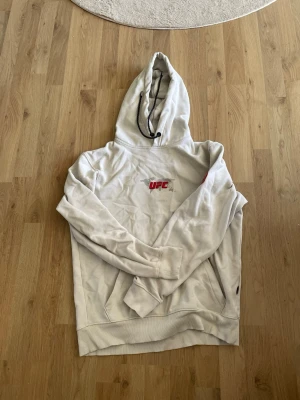 Vit UFC hoodie med tryck - Säljer en vit hoodie från UFC med svart och rött tryck på bröstet och en stor grafisk print på ryggen. Hoodien har huva med dragsko och ribbade muddar. Perfekt för dig som gillar sportig och street-inspirerad stil. Köpt på ett event i Las Vegas också 2025.