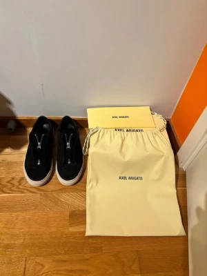 Svarta Axel Arigato sneakers med dragkedja - Snygga svarta sneakers från Axel Arigato med vit sula och dragkedja framtill istället för klassiska snören. Skorna har en stilren och modern design, tillverkade i mocka och levereras med originalpåse och box. Storleken passar till 42-43.