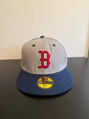 Grå och blå Boston Red Sox keps - Snygg 59FIFTY keps från New Era med Boston Red Sox-logga i rött framtill. Kepsen är grå med mörkblå skärm och detaljer, samt har en klassisk rak form. Perfekt för dig som gillar sportig streetstyle.