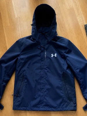 Mörkblå regnjacka Under Armour - Snygg mörkblå regnjacka från Under Armour med justerbar huva och dragkedja framtill. Jackan har två sidofickor med dragkedja och logga på bröstet. Tillverkad i vattenavvisande material, perfekt för regniga dagar.
