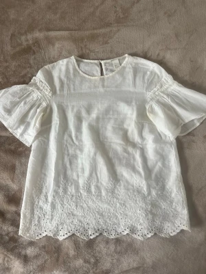 Vit blus med broderade detaljer - Söt vit blus med korta ärmar och rund halsringning. Blusen har broderade blommönster och hålbroderier längs nederkanten samt vågig kant. Tillverkad i ett lätt och luftigt tyg, perfekt för sommaren!!☀️☀️