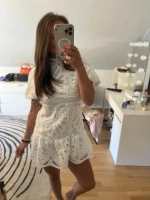 Vit spetsklänning med volang - Superfin vit klänning i spets med korta ärmar och volang nertill. Klänningen har hög hals och ett vackert broderat mönster som ger en romantisk känsla. Perfekt för sommarens alla tillfällen!