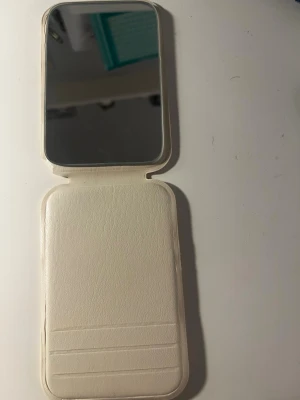 Vit sminkspegel i fodral - Praktisk sminkspegel i rektangulärt format med vitt konstläderfodral. Spegeln är hopfällbar och har plats för kort eller små tillbehör på insidan. Perfekt att ha i väskan för snabba touch-ups när du är på språng.