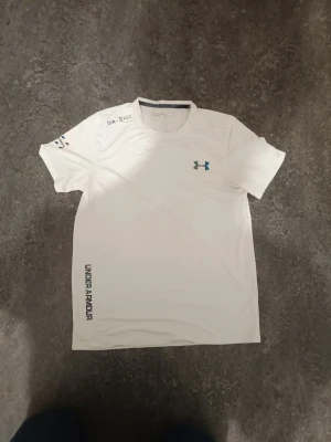  Under Armour T-shirt - Snygg vit t-shirt Under Armour med logga på bröstet och tryck på axel och sida. Tillverkad i lätt och snabbtorkande material, perfekt för träning. Modellen är en klassisk crew neck och har märkningen THREADBORNE för extra komfort.