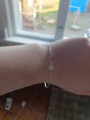 Tunt silverarmband med kors och medaljong - Snyggt och minimalistiskt armband i silverfärg med ett litet kors och en rund medaljong som detaljer. Armbandet har en tunn och stilren design som passar perfekt för dig som gillar diskreta smycken. Perfekt accessoar för att lyfta din outfit. 