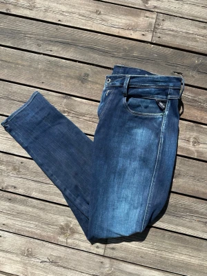 Replay Anbass - Snygga mörkblå slim fit jeans från Replay med klassisk femficksdesign och diskreta slitningar framtill. Jeansen har en något smalare passform och är tillverkade i ett stretchigt denimtyg för extra komfort. Perfekta för en trendig och avslappnad stil. Storlek W34 L32