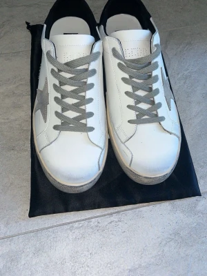 Vita sneakers med grå stjärna - Snygga golden goose skor.Perfekt inför sommaren Knappt använda råkade ha de i en paddelhal så de är lite lite blåa där fram man kan nog tvätta bort det.Stl 45.Fick de av min faster så vet ej om äkthet.Påse tillkommer.Hör av er om fler bilder eller om du vill prata om priset